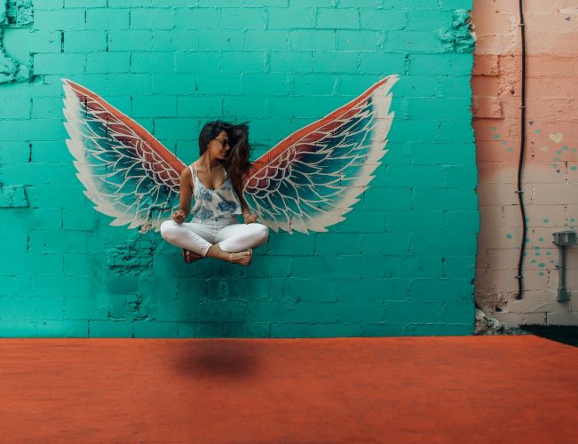 Une femme assise en suspension face à un mur sur lequel sont peints des ailes